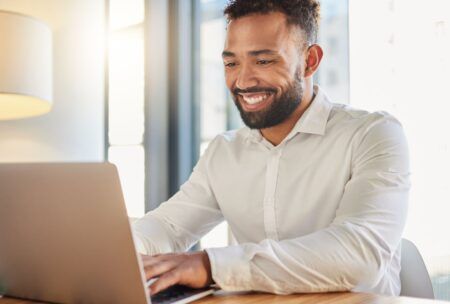 A smiling man sits at a table, working on his laptop, learning his microsoft skills in a productive atmosphere.